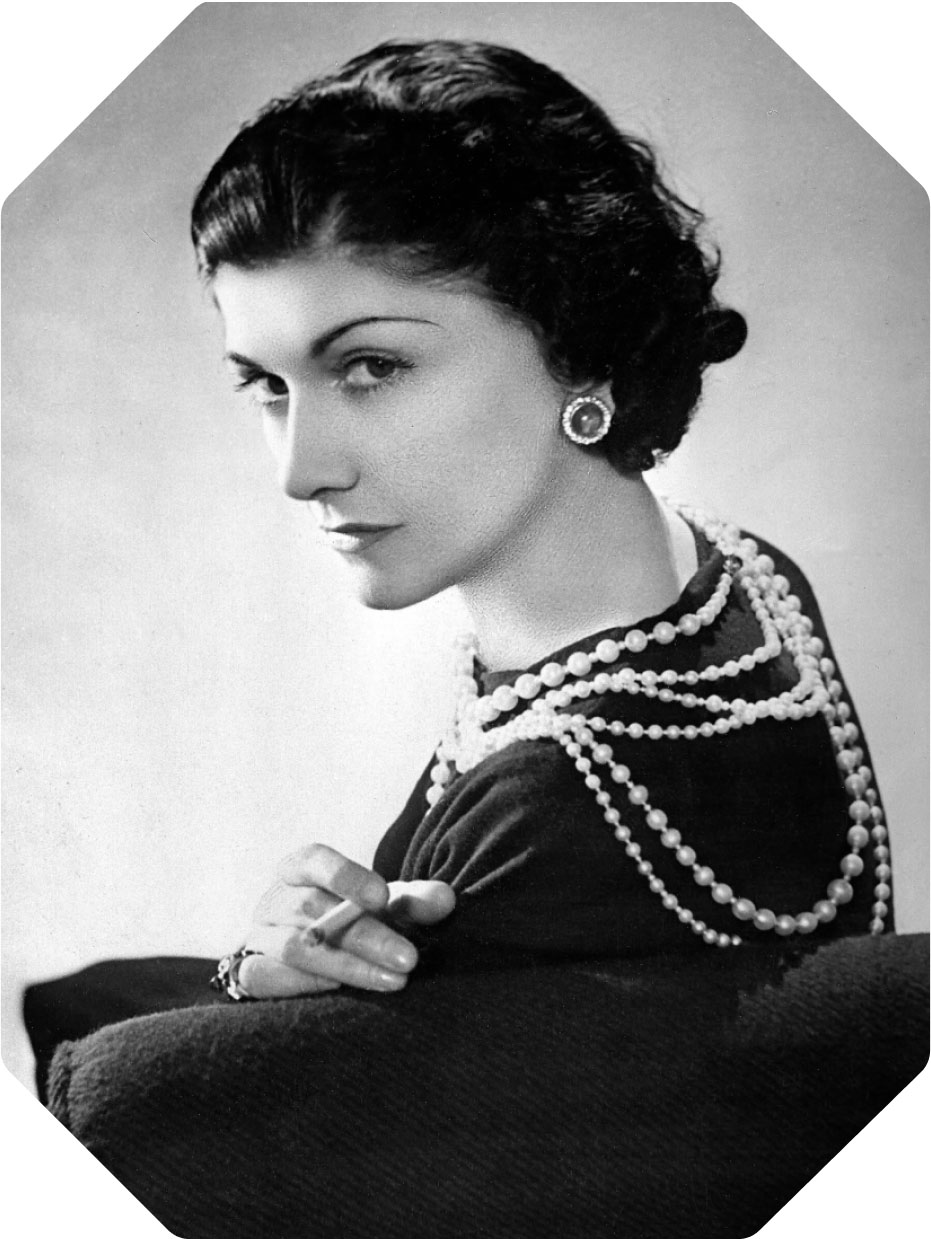 A black and white portrait of Gabrielle Chanel wearing pearls and smoking a cigarette